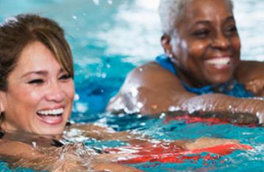 two adult women swimming side by side in an indoor pool, both smiling