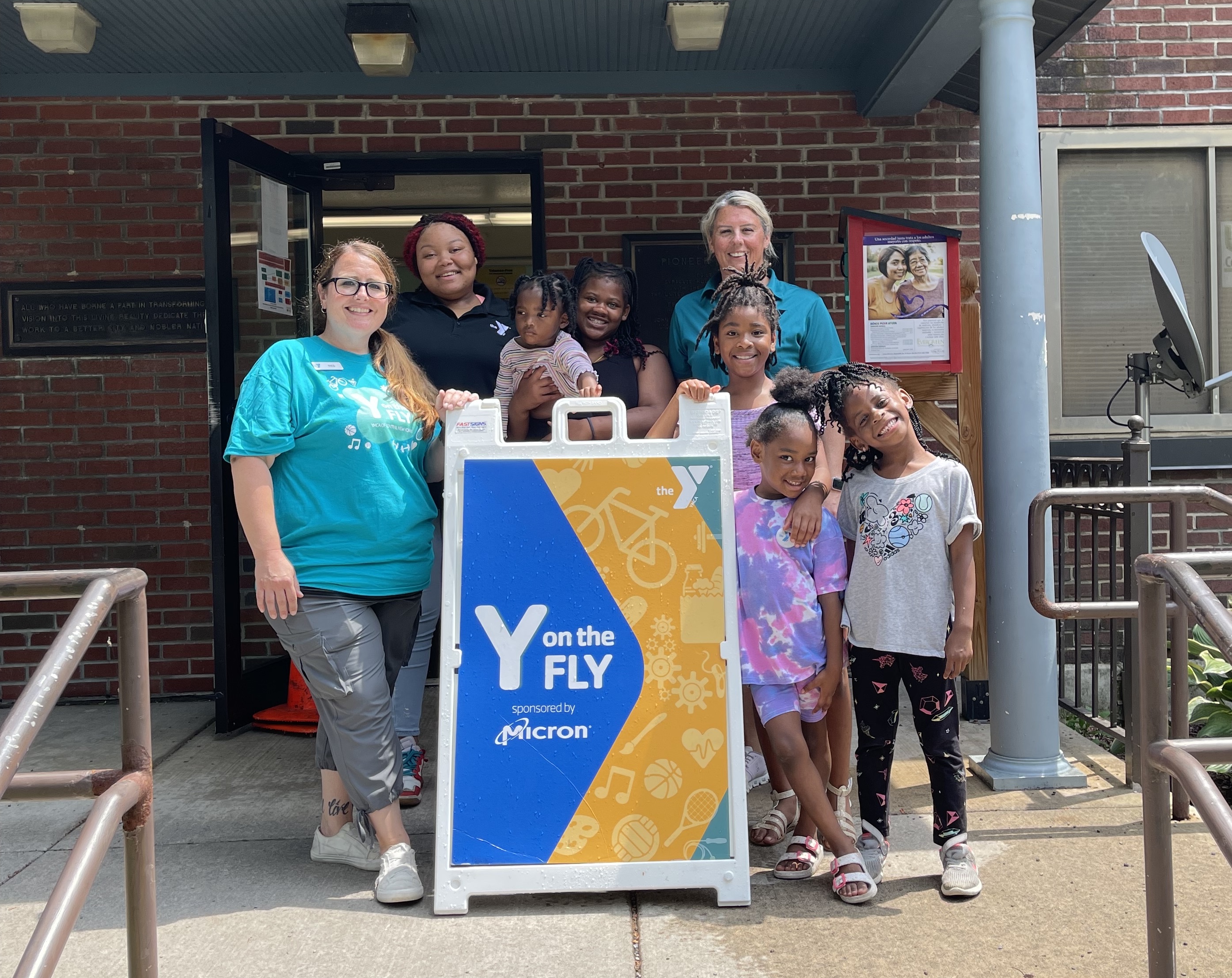 Y staff and kids standing and smiling next to a Y on the Fly sign