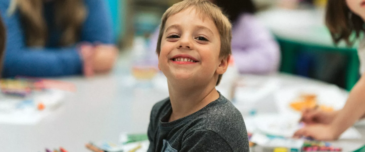 A young child smiles while sitting at a table with a smiling teacher in the background.