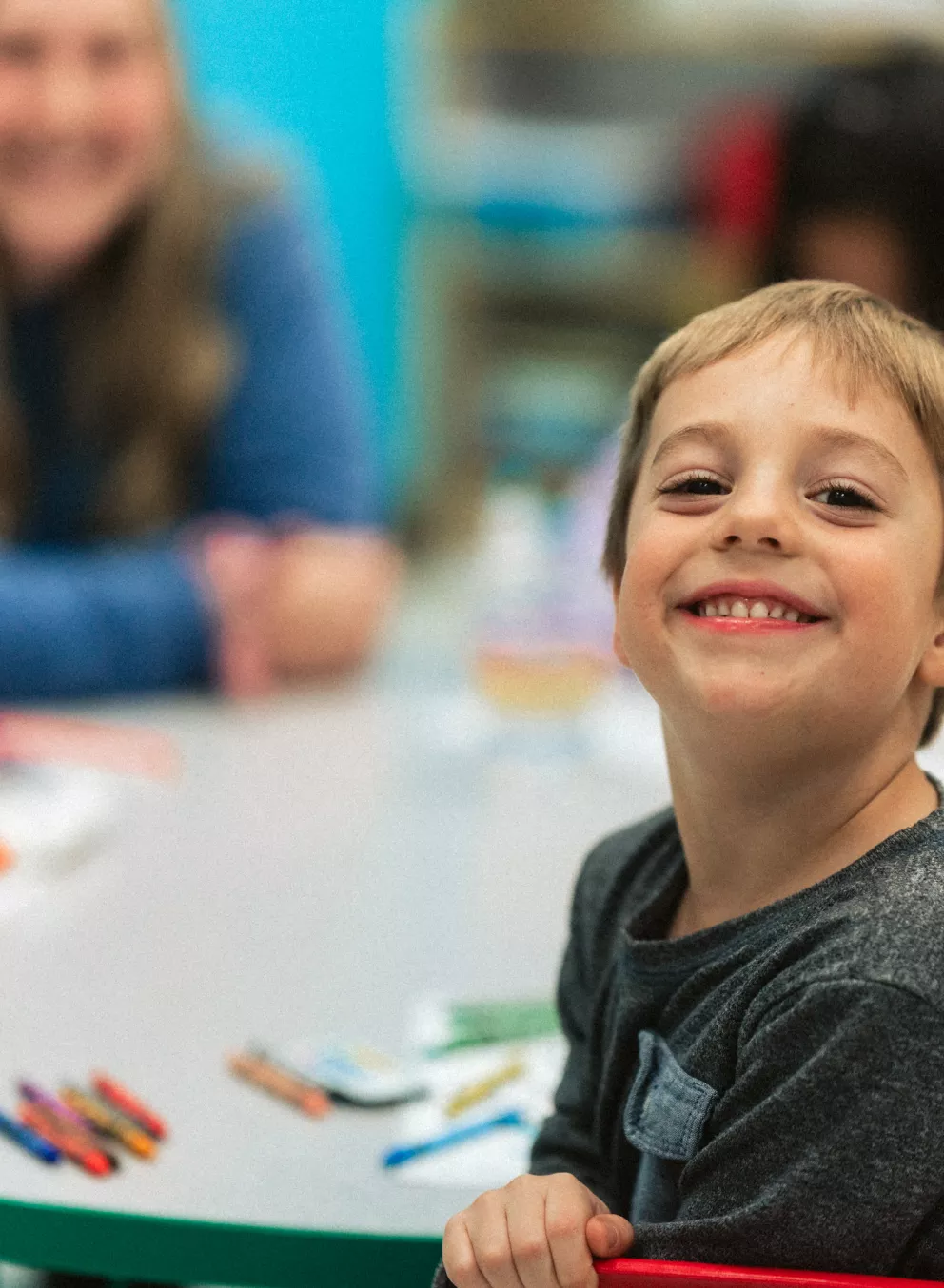 A young child smiles while sitting at a table with a smiling teacher in the background.