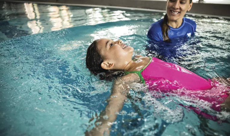 Swim Instructor helps a child in a pink swimsuit learn to float on their back.