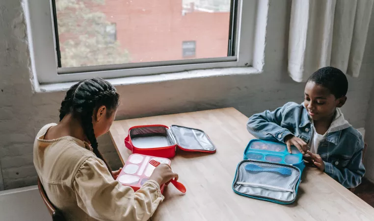 Two kids sitting at a table opening their lunch pals. The food is separated into compartments