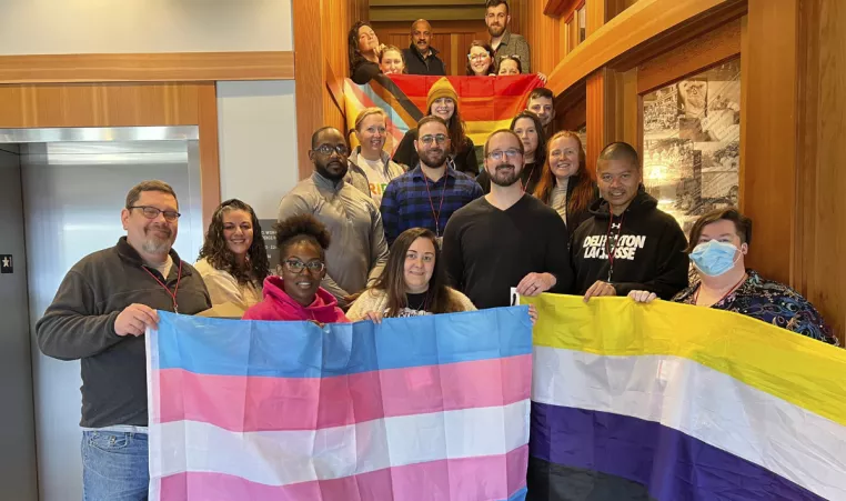A diverse group of people stand on a stair case holding a Transgender Pride flag, a Nonbinary Pride Flag, and a Progress Pride Flag